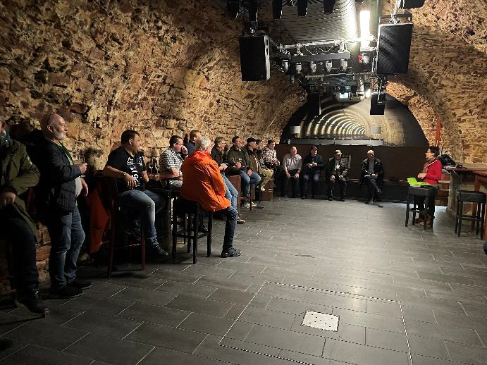 Teilnehmer einer Veranstaltung der Bezirksgruppe Garzweiler sitzen in einem historischen Gewölbekeller mit Bühne und Technik, während eine Vortragende am rechten Bildrand spricht