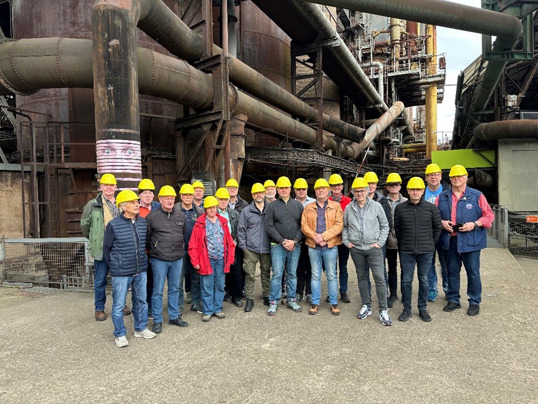 Gruppenfoto mit Besuchern in gelben Helmen vor einer Industrieanlage mit großen Rohrleitungen und Stahlkonstruktionen